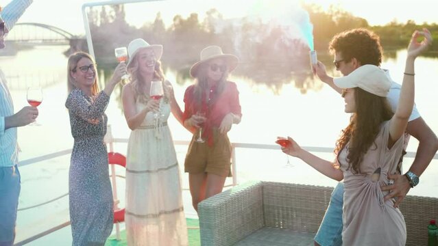 Group of friends dancing, waving color smoke bombs, and toasting enjoying the party on the boat on the river