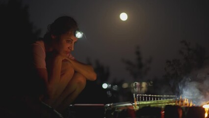 hiker woman with lighting headlamp sitting close to campfire at evening outside wild nature autumn summer vacation. woman cooking meal on fire, BBQ or vegetables on grill. picnic at night