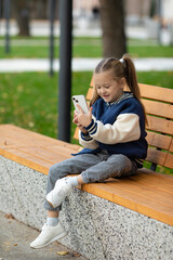 Young Caucasian girl sitting at park and using smart phone. Happy smiling child reading a message on cellphone while sitting on bench in a park. Cute baby surfing net with smartphone