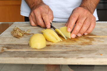 Human hands slicing boiled potato on wooden cutting board with kitchen knife