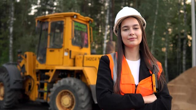 A young woman mine worker is standing in front of a large dump truck or grader. Looking at camera.