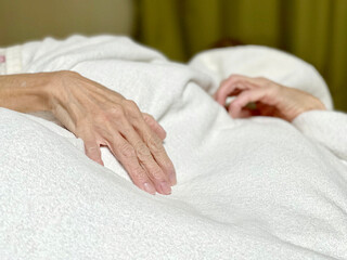 白い布団をかけて寝ている高齢者の手 - obrazy, fototapety, plakaty