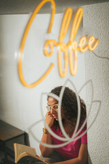 Girl reading book through window in a coffee shop with coffee neon sign