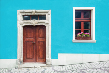 Wooden door home entrance. Window with flower box. Italian architecture background. Vibrant color blue wall facade. Small town house exterior. Street of European city building.