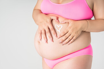 Pregnant woman in pink underwear applying skin stretch mark cream on her belly on gray background, maternity underwear, pregnancy skin care concept