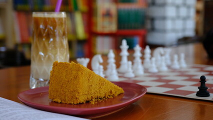a slice of cake, a glass of cold coffee, book and chess board