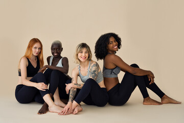 Fototapeta premium Happy diverse young women, 4 multicultural beauty models wearing sportswear sitting at beige background advertising yoga course, gym trainings, team pilates workout, healthy nutrition. Group portrait.