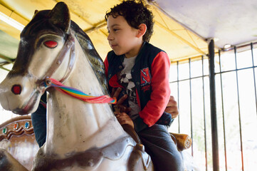 Latino male child riding pretend horse playing with his dad at carousel fair.