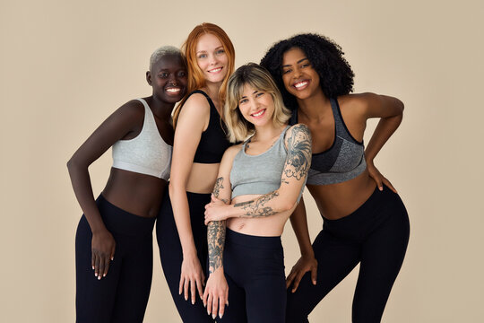 Happy diverse fit sporty young women, 4 multicultural beauty models wearing tops and leggings standing at beige background advertising yoga group gym trainings, team pilates workout. Portrait.