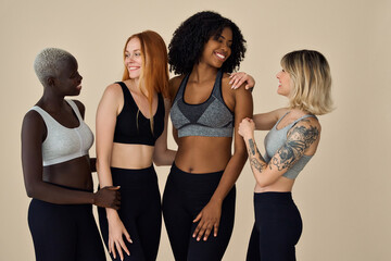 Happy fit sporty diverse young women wear sportswear talking standing at beige background. Multiracial girls advertise sport fitness gym lose weight trainings together, authentic candid shot.