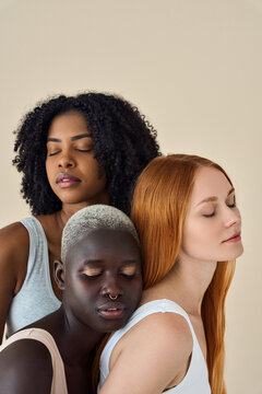 Three pretty diverse young girls bonding with eyes closed isolated on beige background. Tender African and Caucasian women models pretty faces advertising skin care natural beauty. Vertical shot.