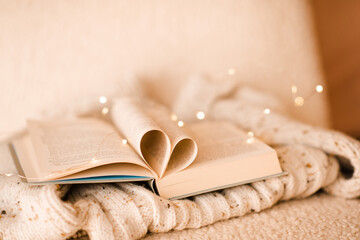 Open paper book with folded pages in heart shape on knitted clothes over Christmas lights in room...