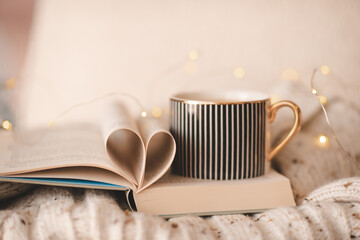 Open paper book with folded pages in heart shape and cup of coffee on knitted clothes over Christmas lights in room close up. Winter cozy home atmosphere.