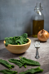 Fresh spinach leaves in a wooden bowl on gray stone table. In the background, oil, onion and a fork, vertical image.