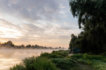 Beautiful summer landscape by the river in the early foggy morning. The tent stands among green...