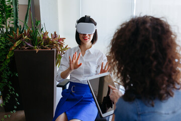 sitting smiling asian woman wearing viewer exploring virtual reality with her left handed colleague...