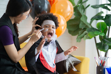 Woman applying Halloween make-up to face of her son at home, sitting together on the sofa in the...
