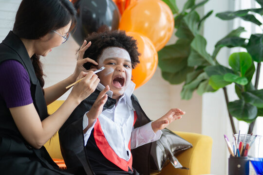 Woman applying Halloween make-up to face of her son at home, sitting together on the sofa in the living room decorated with orange and black balloons, boy acted scary, Halloween celebration