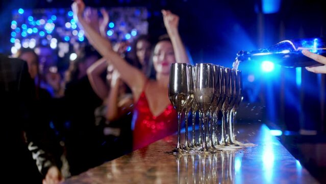 Close-up of a bar riser, the bartender pours champagne. A group of happy people at a party celebrating and drinking champagne in a club, against the backdrop of bright lights.