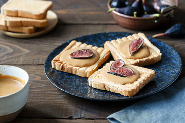 Peanut butter spread on the toasts set on dark blue plate with fig pieces, butter in a bowl, raw figs over wooden background