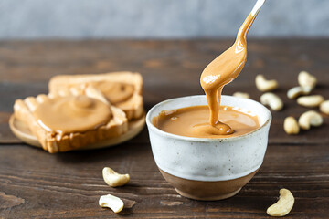 Peanut Butter in a bowl and tea spoon pouring butter to the top, toasts, wooden background, close up