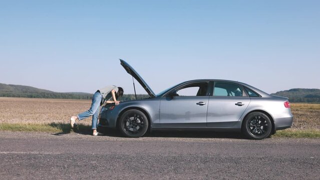 Young woman leaning over looking at car engine breakdown in field, looking frustrated trying to stop cars, slow motion
