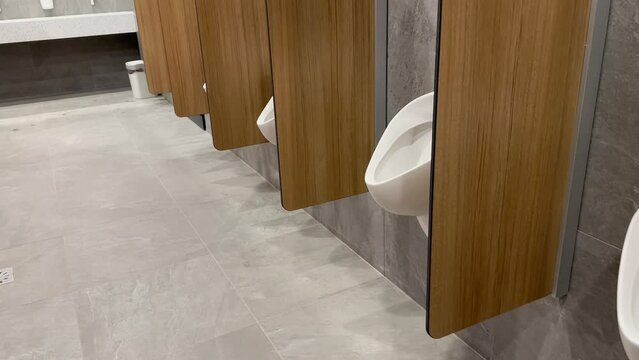 A row of white ceramic urinals are separated by a wooden partition in a men's public toilet in a shopping mall.