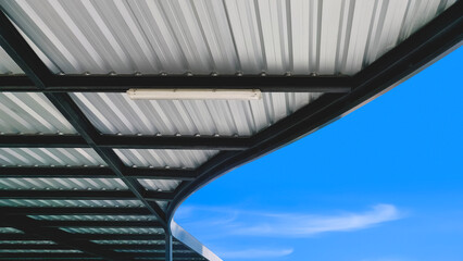 Modern corrugated metal curve roof of covered walkway with black steel roof beam structure against blue sky background, view from below 