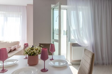 A modern living room combined with a kitchen, in light colors. Dining table with pink tableware