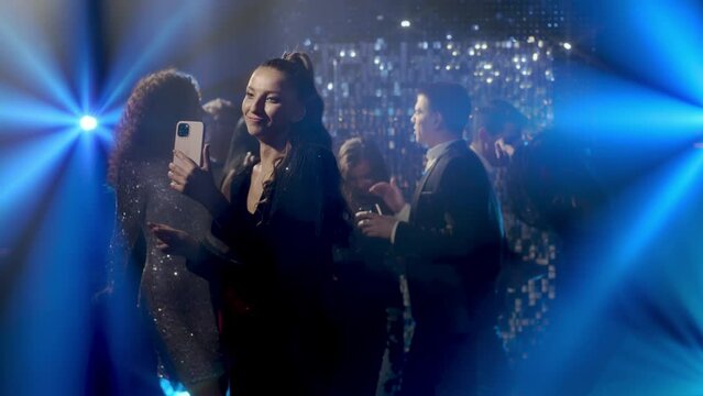 Close-up of a cheerful young woman gesturing in a nightclub. A young female blogger makes a video call on a mobile phone against the background of dancing people or calls her friends.