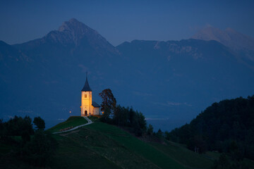 Illuminated The Church of St. Primoz and Felicijan in the village of Jamnik in the Slovenian Alps at night. - obrazy, fototapety, plakaty