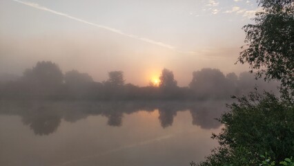 On an early spring morning, the sun rising over the forest of the far side of the river, breaks through the branches of a tree standing on the grassy bank. A colorful sky with feathery clouds. A fog