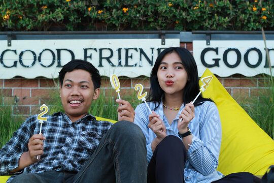 Multiethnic Couple Teen Enjoy Sitting On The Yellow Couch Looking Away And Holding Gold Candle Number 20 24 Waiting New Year Exchange In The House Yard Meadow With Flower And Brick Wall Background