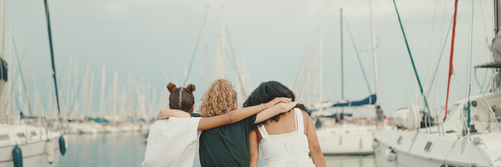 Laughing three girls friends pre-teenage sitting on the waterfront against ships and yachts...
