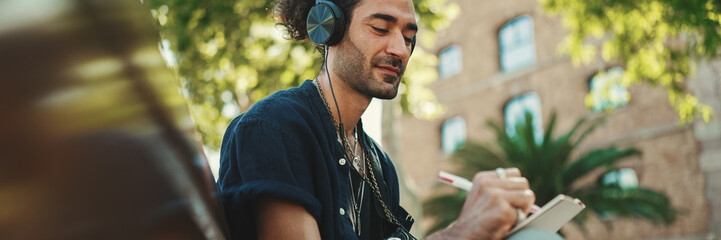 Young italian guy with ponytail and stubble sits with headphones on street bench listens to music...