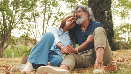 Happy retired couple sitting in the park holding hands and smiling resting on the grass in the park in autumn
