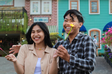 laughing asian couple teen smiling to camera with holding sparklers in the colorful house yard....