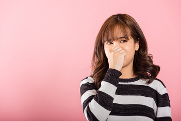 Portrait of an Asian woman, unhappy with a disgusted expression, pinches her nose due to a foul smell. Studio shot isolated on pink, illustrating a healthcare concept. - obrazy, fototapety, plakaty