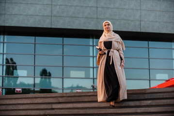 A successful Muslim business woman in hijab walking near the office building. - obrazy, fototapety, plakaty
