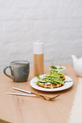 Breakfast on wooden table:zucchini waffles with cottage cheese, avakado and arugula in a white plate, cup of tea. Top view.