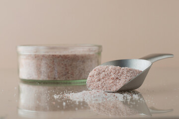 Himalayan pink salt in a glass container with a metal spatula on a beige background on a reflective...
