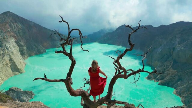 A woman in a red dress stands near a dead tree on the edge of an active volcano Kawah Ijen in the crater of which there is a sulfur lake, East Java, Indonesia. Aerial Drone view 4K.