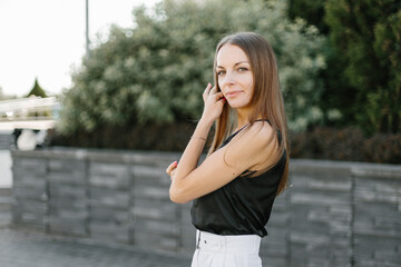 Fototapeta premium Portrait of a girl with long blond hair, wearing a black top, white shorts, poses against a gray wall. Young woman walking through a park with modern architecture and landscape design