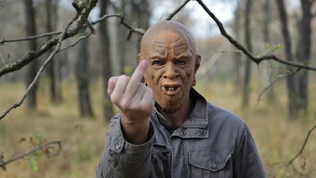 A man in a scary monster mask stands in the forest and shows his middle finger, finger gesturing fuck. A man makes a fuck gesture with his hand against the background of a forest.