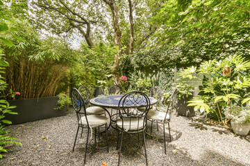 a table and chairs in a backyard setting with trees, plants, and flowers on the side wall behind it