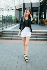 Obraz premium A girl with long blond hair, wearing a black top, white shorts, poses against a background of a gray wall. Young woman walking through a park with modern architecture and landscape design