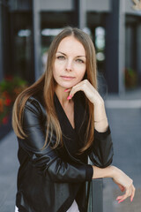 Fototapeta premium A girl with long blond hair, wearing a black top, black jacket and white shorts, poses against the backdrop of modern architecture. Young woman walking in a park with landscape design