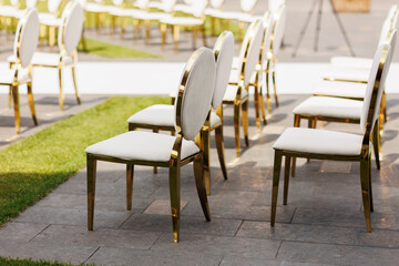 Luxury white-golden chairs on wedding ceremony outdoors. Festive decorations. Empty rows armchairs for guests. Outgoing wedding ceremony in the open air. Decor Studio.