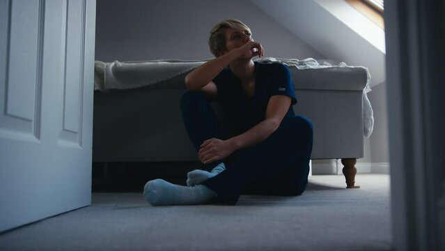 Unhappy woman wearing nurses uniform sitting on bedroom floor at home taking off ID lanyard and questioning career choice - shot in slow motion 
