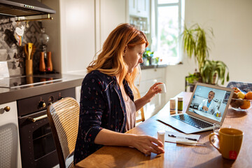 Young woman consulting her doctor on a video call from a laptop in the kitchen at home